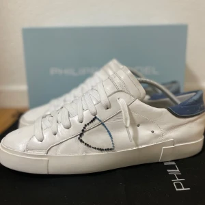 Vita sneakers från Philippe Model - Snygga vita sneakers från Philippe Model med blå detalj på hälen och broderad sköld på sidan. Skorna har rund tå, platt sula och klassisk snörning. Tillverkade i skinn för en lyxig känsla och stilren look. Bra skick! Storlek 43. Box medföljer! Skriv för frågor 💬!