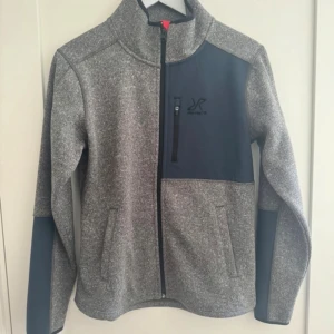 Grå och mörkblå jacka RevolutionRace - Snygg gråmelerad jacka från RevolutionRace i storlek M. Jackan har mörkblå detaljer på bröstet och ärmarna, hel dragkedja framtill och hög krage. Perfekt för dig som gillar outdoor och vill ha en sportig look. RevolutionRace-logga på bröstet och nacken.