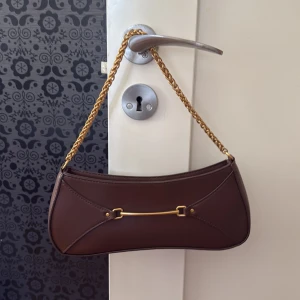 Brun axelväska med guldkedja - Snygg OANVÄND axelväska i skinn med guldfärgad kedja som axelrem och dekorativt gulddetalj framtill. Väskan har en smal, avlång form och stängs med dragkedja. Perfekt för att lyfta din outfit med en elegant touch.