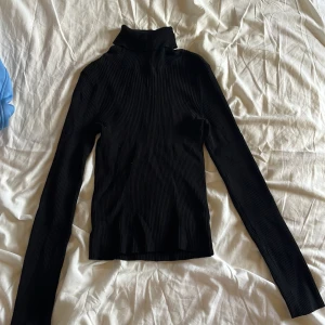 Svart ribbad polotröja - Svart ribbad polotröja med lång ärm och tight passform. Hög krage som ger en clean och stilren look. Perfekt att styla med jeans eller kjol för en enkel men snygg outfit. Materialet känns mjukt och följsamt mot kroppen.