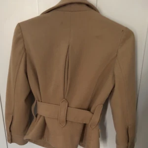 Beige kavajjacka från H&M med bälte - Stilren beige kavajjacka från H&M med breda slag, markerad midja och bälte. Jackan har knappar framtill och fickor med lock, perfekt för dig som gillar klassisk men trendig look. Materialet är mjukt och känns lyxigt.