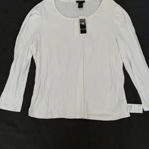 Vit ribbad topp från Design History - Vit långärmad topp från Design History med ribbad struktur och rund halsringning. Dekorativa knappar framtill ger en snygg detalj. Perfekt för dig som gillar en enkel men stilren look. Mjuk och skön känsla, passar till många outfits.