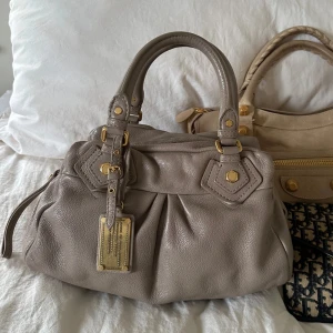 Beige handväska från Marc Jacobs i skinn - Säljer en snygg beige handväska från Marc Jacobs i mjukt skinn med guldiga metalldetaljer och dekorativ metallbricka framtill. Väskan har dubbla handtag och veckad design som ger en cool och lyxig vibe. Perfekt för dig som gillar klassiska men trendiga accessoarer.