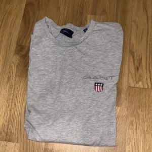 Grå t-shirt från GANT med broderad logo - Snygg grå t-shirt från GANT med klassisk rund hals och korta ärmar. På bröstet finns en broderad GANT-logo och ett litet sköldmärke i rött, vitt och blått. T-shirten är i mjuk bomull och har en clean, stilren look som funkar till allt.