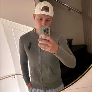 Grå stickad kofta från Polo Ralph Lauren - Säljer en grå stickad kofta från Polo Ralph Lauren med dragkedja, två fickor och stor mörkblå logga på bröstet. Snygg ribbad krage och ärmslut samt en broderad siffra 3 på ena ärmen. Perfekt för lager-på-lager-stil.