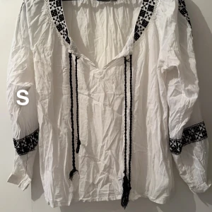 Vit blus med broderi och snörning - Vit långärmad blus med svart broderi på krage och ärmslut. Skjortan har v-ringning och långa svarta snören framtill. Luftig passform och tillverkad i bomull, perfekt för en avslappnad och trendig look.🌸 gulare under armhålorna. Storlek s 