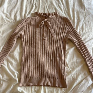 Ljusrosa ribbad stickad tröja med rosett - Supersöt ljusrosa stickad tröja med ribbat mönster och lång ärm. Hög hals med volangkant och en fin rosett framtill som ger en extra detalj. Perfekt för dig som gillar en stilren men ändå gullig look 🩷