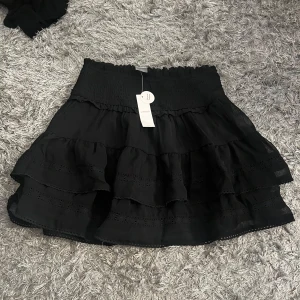 Svart volangkjol från Lindex - Supersöt svart kortkjol från Lindex med flera lager volanger och smock i midjan. Kjolen har fina spetsdetaljer och är perfekt för dig som gillar en flowy och trendig stil. Lätt och luftig, passar till många olika looks.