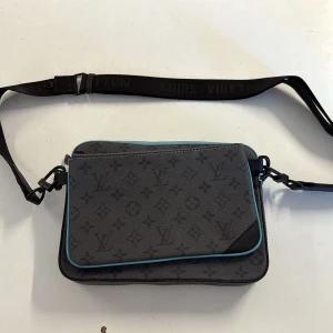 Louis Vuitton axelväska monogram grå - Snygg axelväska från Louis Vuitton i grått skinn med klassiskt monogrammönster och svarta detaljer. Väskan har turkosa kanter, justerbar axelrem med logga och flera fack. Perfekt för dig som vill ha en stilren och lyxig accessoar med ikonisk design.