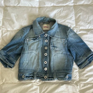 Blå kortärmad croppad jeansjacka från ONLY - Snygg croppad kortärmad jeansjacka från ONLY i ljusblå denim. Jackan har korta ärmar, stora silvriga knappar framtill och broderade blommor på bröstet och ryggen. Klassisk krage och coola detaljer vid midjan. Perfekt för dig som vill ha en trendig och unik look. Storlek M. Bra skick!