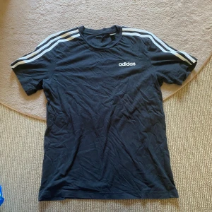 Adidas t-shirt - Svart t-shirt från Adidas i bomull med klassiska vita ränder längs axlarna och Adidas-logga på bröstet. Kortärmad och normal passform, perfekt för en sportig look eller chill vardagsstil!🙌🏻