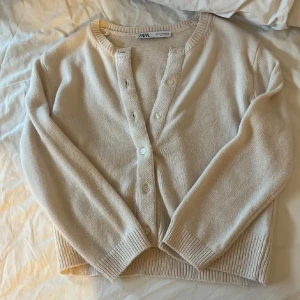 Beige kofta från Zara i stickad bomull - Supermjuk beige kofta från Zara i stickad bomull. Klassisk modell med rund hals, ribbade muddar och knappar framtill. Perfekt att slänga över en t-shirt eller linne för en chill och stilren look.liten i modell