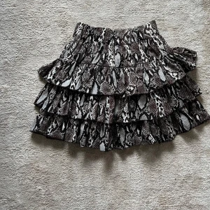 Omlott kjol  - Supersnygg kort volangkjol med svart, vit och brun ormmönstrad design. Kjolen har flera lager volanger som ger extra volym och en cool look. Perfekt för dig som vill sticka ut med ett djurmönster och gillar trendiga detaljer. Storlek 12Y men passar S och Xs