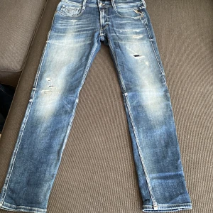 Replay Anbass blå jeans herr 32 - Säljer ett par Replay Anbass jeans i klassisk blå tvätt med slitningar och snygga detaljer. Jeansen har smal passform, fem fickor och tydliga slitna partier framtill för en cool look. Perfekta för dig som gillar en modern och avslappnad stil.