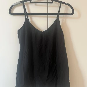 Svart linne med smala axelband - Enkel och stilren svart linnetopp med smala, justerbara axelband och v-ringad halsringning. Perfekt att matcha med jeans eller kjol för en clean look. Tunt och mjukt material som känns skönt mot huden.