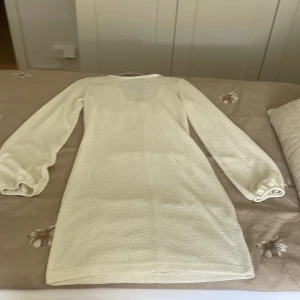 Vit kortklänning med svart rosett - Supersöt vit kortklänning med långärmade puffärmar och en stor svart rosett i satin bak i nacken. Klänningen har en lätt textur i tyget och detaljer med paljetter på rosetten. Perfekt för dig som vill sticka ut med en chic och lekfull look.