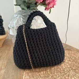 Svart stickad handväska med kedja - Unik svart handväska i grovstickad bomull med snygg guldig kedja som axelrem. Väskan har rundad form och två handtag upptill, perfekt för dig som gillar en personlig och trendig accessoar. Enkel att matcha med olika outfits och rymmer det viktigaste.