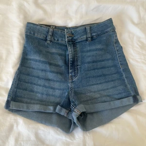 Högmidjade jeansshorts från H&M - Blåa högmidjade jeansshorts från Divided med klassisk femficksdesign, uppvikta benslut och bälteshällor. Perfekta för varma dagar och enkel att matcha med allt. Snygg och tidlös modell som alltid funkar