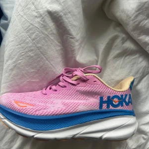 Hoka rosa sneakers med blå sula - Snygga sneakers från Hoka i en fräsch rosa färg med blå och vit tjock sula. Ovandelen är i mesh-liknande syntetmaterial med orange detaljer och beige insida. Skorna har snörning och en sportig vibe, perfekta för dig som gillar färg och komfort.
