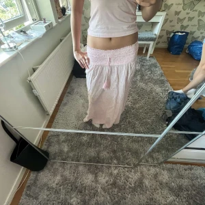 Ljusrosa långkjol - Supersöt ljusrosa långkjol med bred smock i midjan och dekorativt knyt med tofsar framtill. Super snygg💕💕