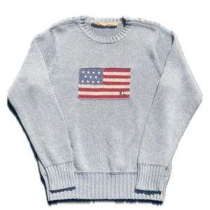 Grå stickad tröja Ralph Lauren - Grå stickad tröja från Ralph Lauren med amerikansk flagga broderad på bröstet och RL-logga. Klassisk rund halsringning och ribbade muddar vid ärmslut och nederkant.