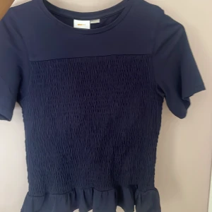 Mörkblå topp med volangkant - Supersöt mörkblå topp med korta ärmar och smockad front och rygg. Toppen har en volangkant längst ner som ger en extra touch. Storlek 146/152 men töjbar och sitter bra på mig som är 174! 