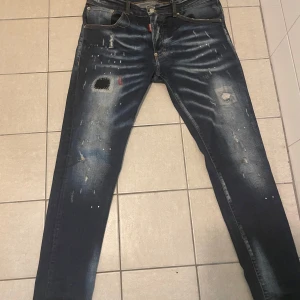 Mörkblå Dsquared2 ICON jeans  - Säljer ett par mörkblå Dsquared2 ICON jeans med coola slitningar, patchar och färgstänk. Jeansen har en smal passform, klassisk femficksdesign och tydlig röd ICON-label bak i midjan. Perfekta för dig som gillar streetstyle och detaljer.