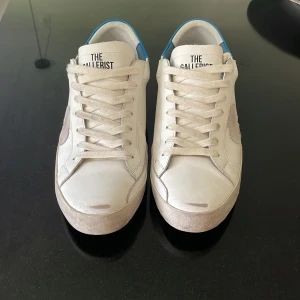 Vita sneakers från The Gallerist strlk 43 - Snygga vita sneakers från The Gallerist, storlek 43, innermått 27,5 cm. Blå detalj på hälen, ”distressed leather” enligt design och märke i mocka på sidan. Knappt använda, nästan i nyskick. Nypris 2 250kr. Perfekta för dig som gillar stilrena men unika detaljer.