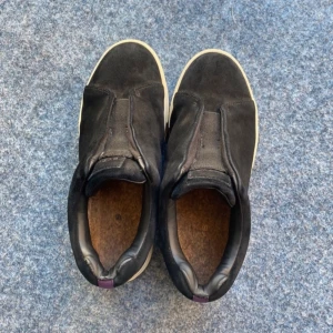 Etys skor till salu! - Stilrena svarta sneakers från ETYS i mjuk mocka med beige sula och lila detalj baktill. Skorna har en platt sula och rund tå, perfekt för en clean och modern look. Innersulan är i kork för extra komfort.