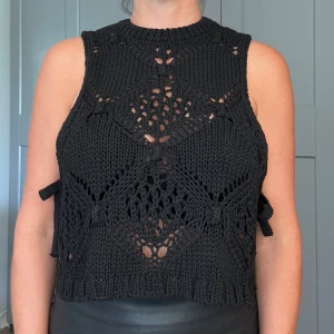 Svart stickad ärmlös topp från Zara - Svart stickad topp från Zara med grovt virkat mönster och höghalsad design. Toppen är ärmlös, croppad och har snygga knytband i sidorna som ger en cool detalj. Perfekt att styla över ett linne eller bikini för en trendig look.