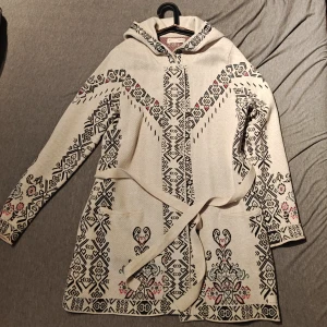 Odd Molly beige cardigan jacka mönstrad - Säljer en beige cardigan jacka från Odd Molly med svart och rosa broderat mönster, huva och knytband i midjan. Jackan är i 100% bomull och har långa ärmar samt två fickor framtill. Perfekt för dig som gillar unika detaljer och boho-vibe.
