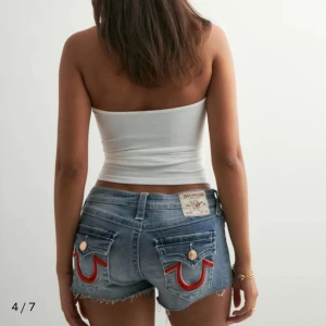True Religion jeansshorts - Snygga blå jeansshorts från True Religion med ikoniska röda sömmar på bakfickorna och råa kanter. Klassisk femficksmodell, låg midja och guldiga knappar. Perfekta för sommaren och ger en cool vibe till din outfit. Nypris är 850.
