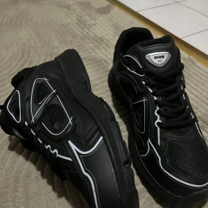 Svarta Dior B30 sneakers - Snygga svarta Dior B30 sneakers med vita detaljer och reflekterande linjer. Skorna har en chunky sula, meshpaneler och partier i syntet och skinn. Klassisk snörning och tydlig Dior-logga på plös och sula. Perfekta för dig som gillar statement sneakers.