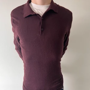 Vinröd merinoull pikétröja Dressmann - Mörk vinröd långärmad pikétröja från Dressmann i mjuk merinoull. Klassisk krage med två knappar framtill och smal passform. Perfekt för dig som gillar stilrena och bekväma plagg med en elegant touch.