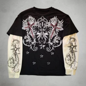 Affliction Longsleeve - Unik svart långärmad tröja med kontrasterande vita ärmar och detaljerat tryck med stjärnor, kors och ornament i vitt, rött och svart. Ärmarna har coola grafiska motiv och tröjan har en edgy streetstyle-vibe. Perfekt för dig som gillar statement-plagg.