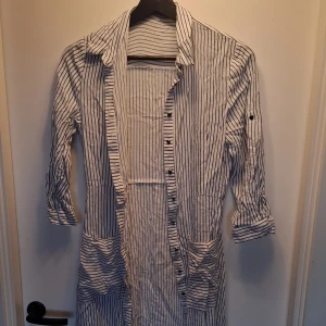 Randig långklänning från Zara Basic - Långklänning från Zara Basic i vitt med smala blå ränder. Klänningen har knappar hela vägen framtill, stora fickor och trekvartsärmar som kan rullas upp. Snygg krage och avslappnad passform, perfekt för en chill sommarlook.