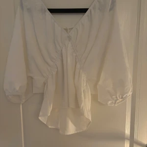 Vit blus med knyt och volang - Supersöt vit blus  med knyt i ryggen och v-ringning. Blusen har trekvartsärmar med resår och volangig nederkant. Materialet är lätt och har ett subtilt mönster i tyget som ger en lyxig känsla. Perfekt till jeans eller kjol för en fräsch look, den är lite nopprig men inget som syns på nära eller långt håll! Nypris är 400 kr 
