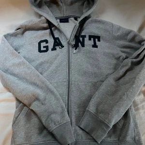 Grå hoodie från GANT med dragkedja - Snygg grå hoodie från GANT med svart logga över bröstet. Tröjan har huva med snörning, hel dragkedja och två fickor framtill. Tillverkad i mjuk bomullsmix med insida i fleece för extra komfort. Perfekt för chill dagar eller när du vill ha en clean look.