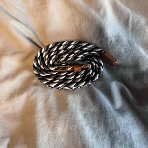 Randigt flätat bälte med läderdetaljer - Snyggt flätat bälte i svart och vitt med randigt mönster. Bältet har bruna läderdetaljer vid ändarna och är flexibelt i materialet. Perfekt accessoar för att lyfta din outfit med en cool och stilren touch.