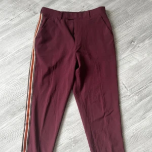 Vinröda byxor med revärer - Snygga vinröda byxor med coola revärer i orange, rött och vitt längs sidorna. Byxorna har fickor fram och bak samt hällor för bälte. Perfekta för dig som vill sticka ut lite extra.