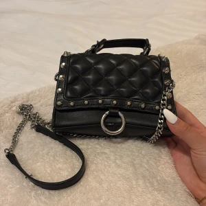 Svart quiltad axelväska med kedja - Snygg svart axelväska i quiltat skinn med silverfärgade nitar och kedjedetaljer. Väskan har ett handtag upptill, avtagbar axelrem i kedja och en stor silverring framtill. Perfekt för att lyfta vilken outfit som helst.