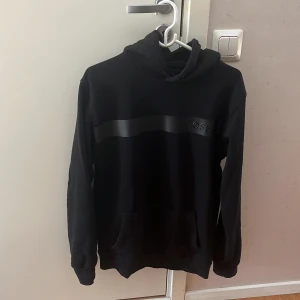 Svart hoodie från BOSS med logga - Svart hoodie från BOSS med stilren svart logga och bred rand över bröstet. Tröjan har huva, känguruficka och långa ärmar. Perfekt för dig som gillar en clean och klassisk streetstil.