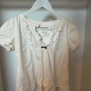 Vit blus med volang från Odd Molly - Supersöt vit blus från Odd Molly med korta puffärmar, volangdetaljer längs v-ringningen och en liten svart rosett framtill. Har en liten fläck. 