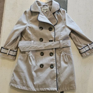 Beige trenchcoat från Milkshake till barn - Stilren beige trenchcoat från Milkshake med svarta detaljer längs krage och ärmslut. Jackan har dubbelknäppning, bälte i midjan och klassisk krage. Perfekt för dig som vill ha en snygg och tidlös look under vår och höst.