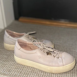Beige sneakers från Dasia i skinn - Snygga beige sneakers från Dasia i mjukt skinn med platt, vit sula och rund tå. Skorna har snörning med guldfärgade detaljer och en liten metallcharm på ena snöret. 