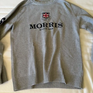 Grå stickad tröja från Morris - Snygg grå stickad tröja från Morris med broderad logga och brittisk flagga på bröstet. Tröjan har ribbade muddar vid ärmslut och nederkant samt rund hals. Perfekt för dig som gillar klassisk och stilren design.