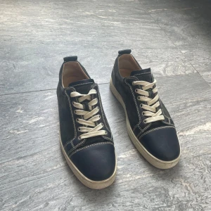 Christian Louboutin skor - Snygga svarta sneakers från Christian Louboutin med kontrasterande vita sömmar och vita snören. Ovandel i mocka och skinn, klassisk rund tå och ikoniska röda sulor undertill. Perfekta för dig som vill sticka ut med en lyxig touch.
