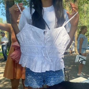 Vit volangig linnetopp med knyt - Säljer en söt vit linnetopp med smala axelband, volangkant nertill och knytning framtill. Toppen har en lätt rynkad effekt och är perfekt för varma dagar. Enkel och fräsch stil som passar till jeansshorts eller kjol.