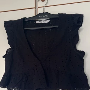 Svart blus med volang från NA-KD - Supersöt svart blus från NA-KD i storlek 36. Den har broderade hål, v-ringning och volangdetaljer längs ärmar och nederkant. Ärmlös modell med romantisk känsla, perfekt till jeans eller kjol för en trendig look.