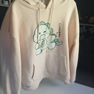 Beige hoodie med söt dinosaurieprint - Supermysig beige hoodie med en gullig mintgrön dinosaurie som dricker bubble tea på framsidan, knappt använt den max 3 gånger jätteskön men säljer pga av att den är för stor för mig.